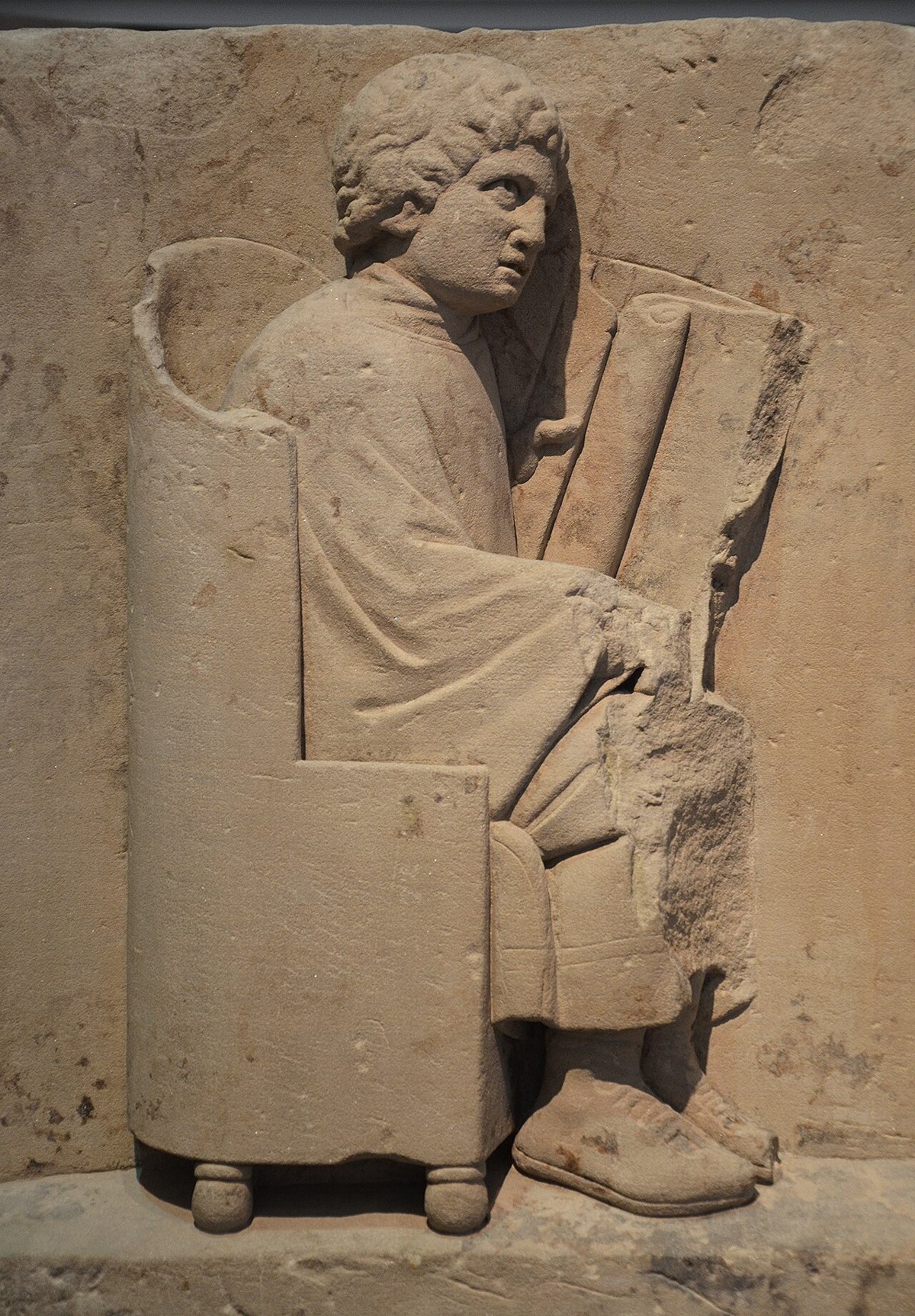 Roman sandstone relief of a young reader seated in a high-backed chair, holding an unrolled scroll